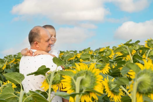 Father's day concept. A little boy in his father's arms, in white clothes, gently hugs and cuddles, against the blue sky, in a field with yellow sunflowers. A symbol of peace. Symbol of Ukraine. Close-up. copy space.