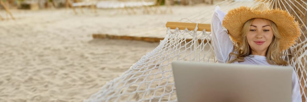 Work remotely. Side view of a young happy female freelancer in a straw hat working on a laptop while relaxing in a hammock on the beach at sunset. Remote work during vacation.