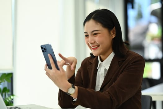 Happy female entrepreneur in luxury suit using smartphone during break from work at office.