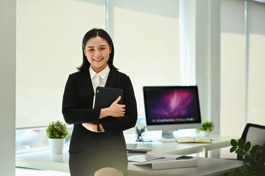 Portrait of confident millennial female manager in black suit standing with arms crossed and smiling at camera.
