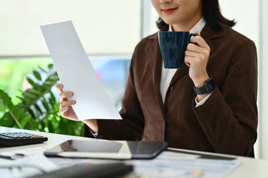 Pleasant female manager drinking coffee and analyzing sales statistics document at working desk.