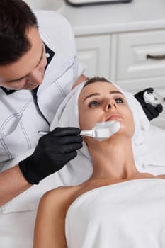 male beautician applies the mask to the face of beautiful woman in the spa salon. facial skin care concept.