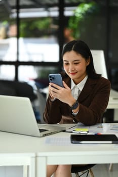 Charming asian woman entrepreneur sitting in bright home office and using smartphone. Business, technology and people.