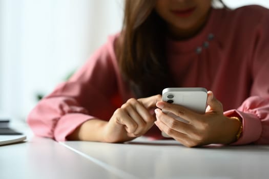 Cropped image of young woman using smartphone at office desk, checking social media, communicating online.