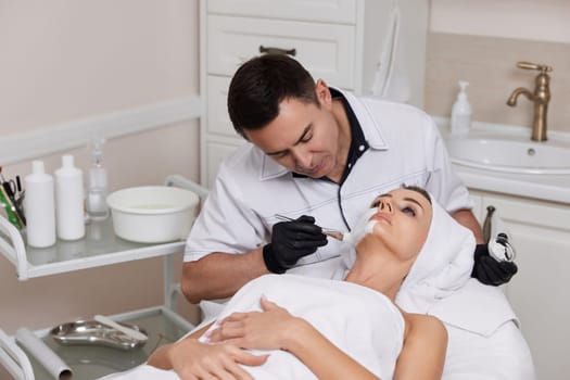 male beautician applies the mask to the face of beautiful woman in the spa salon. facial skin care concept.