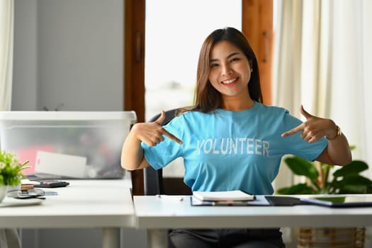 Positive young woman, volunteer pointing fingers at blue t-shirt and smiling to camera.