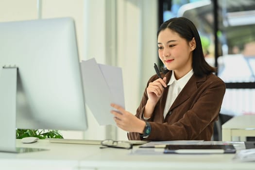 Millennial woman entrepreneur holding documents and watching online webinar on computer screen.