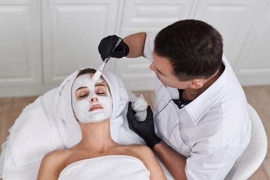 male beautician applies the mask to the face of beautiful woman in the spa salon. facial skin care concept.