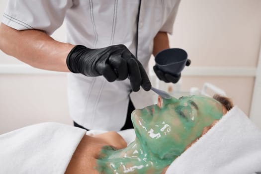 beautician applies green alginate mask to the face of beautiful woman in the spa salon. spa treatments