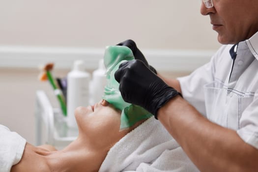 male beautician removing green alginate mask from face of beautiful woman in the spa salon. spa treatments