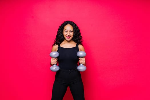 Smiling african american female doing hand exercises with dumbbells on a red studio background.