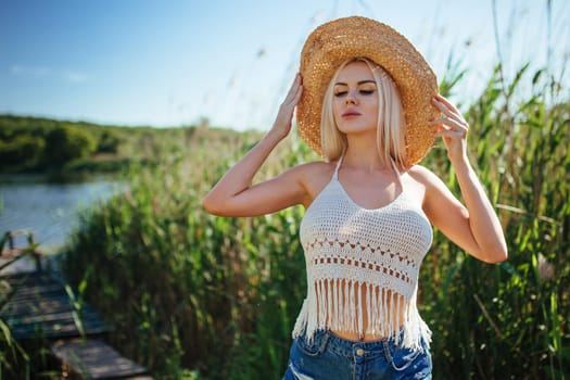 Beautiful model girl posing on the field enjoying nature outdoors in a straw hat. Beauty blond young woman with long straight blond hair close up portrait.