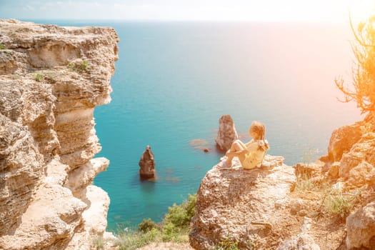 Happy girl perched atop a high rock above the sea, wearing a yellow jumpsuit and braided hair, signifying the concept of summer vacation at the beach