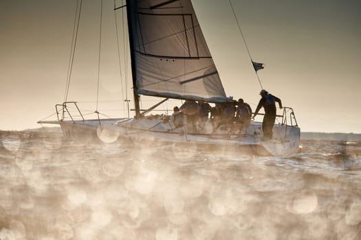 Russia, St.Petersburg, 07 July 2023: The view through the spray of how the sailboat is heeling at sunset, boat roll, splashes shine in the sun, sailors on the board. High quality photo