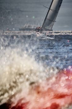 Russia, St.Petersburg, 07 July 2023: The view through the spray of how the sailboat is heeling at sunset, boat roll, splashes shine in the sun, sailors on the board. High quality photo