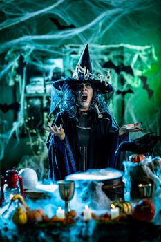 Witch with awfully face in creepy surroundings and smoky green background talks magic words to bones around her hat, above boiling cauldron. Halloween concept.