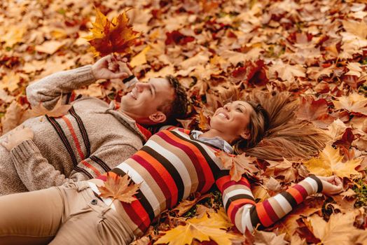 Beautiful young couple in sunny forest in autumn colors. They are lying on the ground covered with fallen leaves and enjoying. 