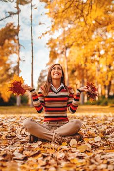 Cute young woman enjoying in sunny forest in autumn colors. She is holding golden yellow fallen leaves and having fun. 