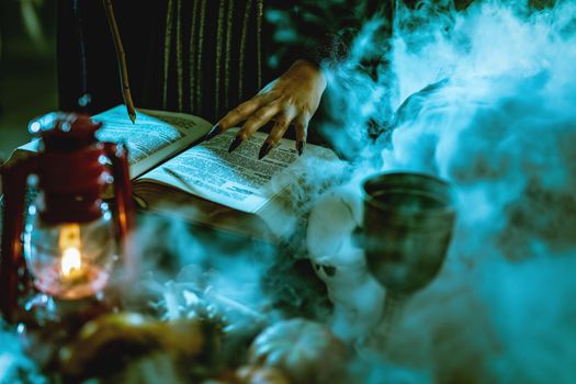 Close-up of a witch's hand with pointed black nails on smoky book of magic recipe and with a magic wand.