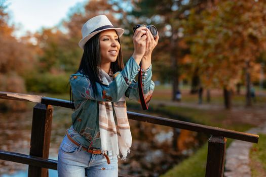 Happy woman photographer holding professional digital camera and taking photo near the lake in autumn park.  