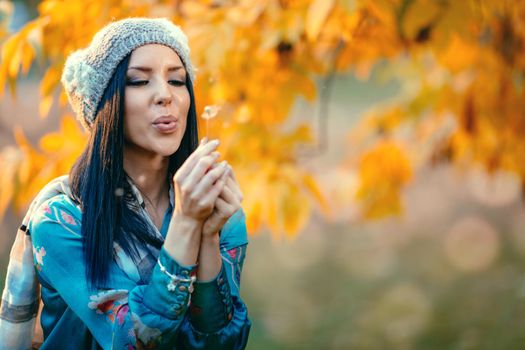 Portrait of young positive beautiful smiling young woman blowing dandelion in autumn park.