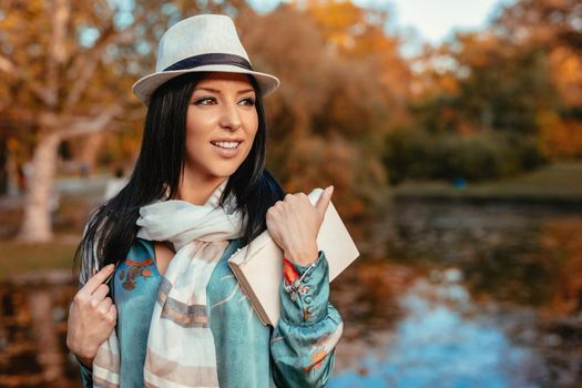 Young happy woman enjoying in a beautiful autumn golden day with a book, near the little lake in the park.