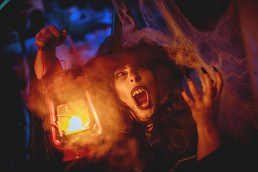 Portrait of witch with awfully face and lighted lantern in her hand in creepy foggy surroundings sends evil.