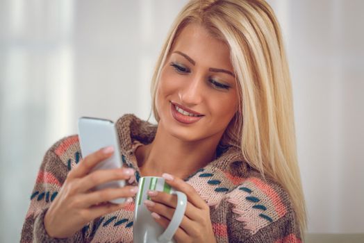 Beautiful young woman in a wool sweater, looking at mobile phone that holds in her hand, and in the other hand holding a cup of tea.