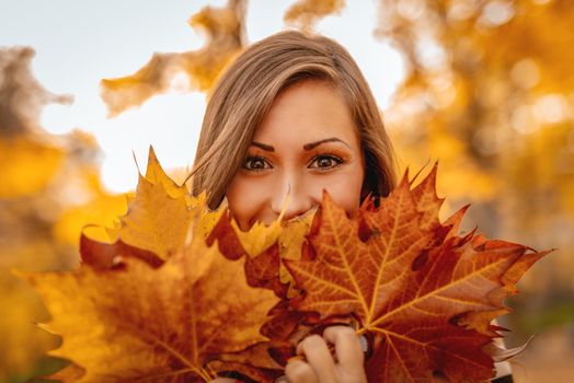 Cute young woman enjoying in sunny forest in autumn colors. She is holding many fallen leaves and looking at camera behind them.