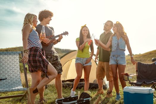 Young people have a good time in camp in nature. They're resting, laughing and singing with music from ukulele, happy to be together.