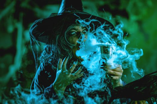 Portrait of witch with awfully face in creepy surroundings and smoky green background drinks magic potion from the goblet.