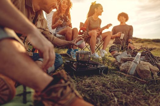 Young people have a good time in camp in nature. They're resting, laughing and singing with music from ukulele, happy to be together.