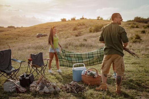 The young couple is preparing for camping. They're putting a picnic blanket on a suitable place in a sunset.