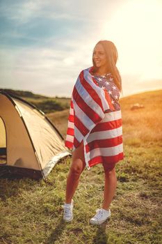Happy young woman enjoys a sunny day in nature. She's wrapped in american flag in front a campsite tent. 