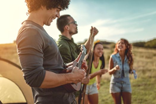 Young people have a good time in camp in nature. They're resting, laughing and singing with music from ukulele, happy to be together.