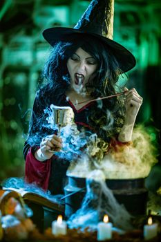 Young witch with seriously face in creepy surroundings and smoky green background holds a goblet with magic potion. 
