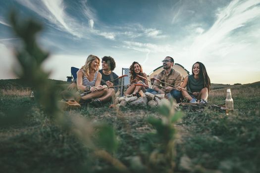 Happy young friends enjoy a sunny day at the mountain. They're laughing and roasting sausages on sticks over a campfire near tent.