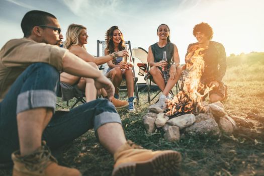 Happy young friends enjoy a sunny day at the mountain. They're laughing and toasting with beer bottles by the bonfire near tent.