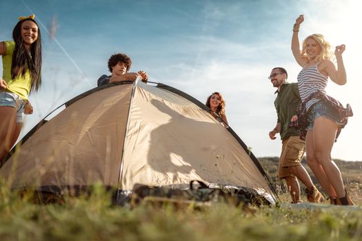 The young happy friends are preparing for camping. They're installing a tent on a suitable place in a meadow and their girlfriends are helping them. Girls dance in nature. 