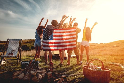 Happy young friends enjoy a sunny day in nature. They're looking at sun holding american flag and greeting, happy to be together.