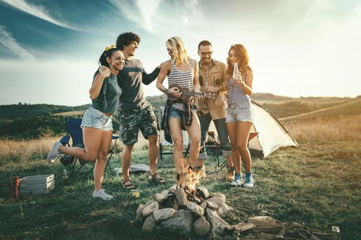 Happy young friends enjoy a sunny day at the mountain. They're laughing and singing with music from ukulele by the bonfire near tent.