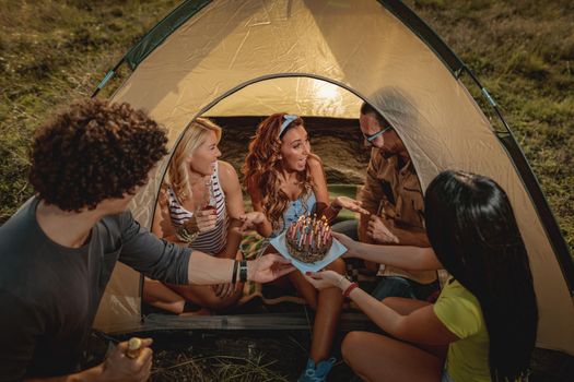 Young people have a good time in camp in nature. They're celebrating a birthday, laughing and greeting to their friend with birthday cake, happy to be together.