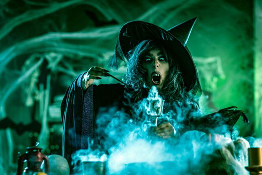 Portrait of witch with awfully face in creepy surroundings and smoky green background drinks magic potion from the goblet.