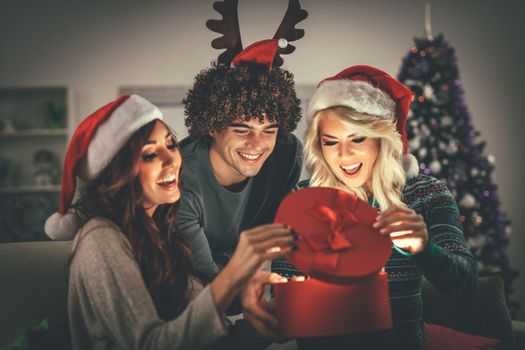 Cheerful beautiful young friends enjoying Christmas at home, opening gift and surprisingly looking what is inside.
