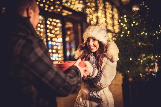 Young beautiful cheerful couple celebrating Christmas in the city street and giving gift to each other. 