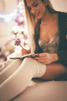 Cute young smiling woman sitting on the bed and making Christmas wish list in a her notes. 