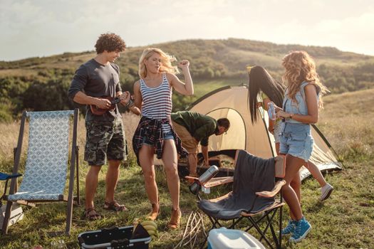 Young people have a good time in camp in nature. They're resting, laughing and singing with music from ukulele, happy to be together.