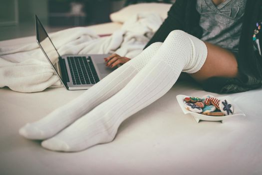 Close-up of the young woman's legs sitting on the bed near colorful cookies on the little plate, making Christmas wish list on her laptop. 