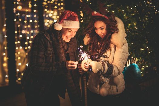 Cute cheerful couple with sparklers enjoying at the Christmas eve in the city street with a lot of holiday's lights.