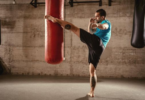 Muscular man punching with leg a boxing bag on cross fit training at the gym.